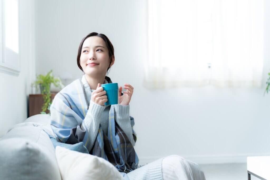 厚手のニットでマグカップを持ちソファでくつろぐ日本人女性、明るい室内